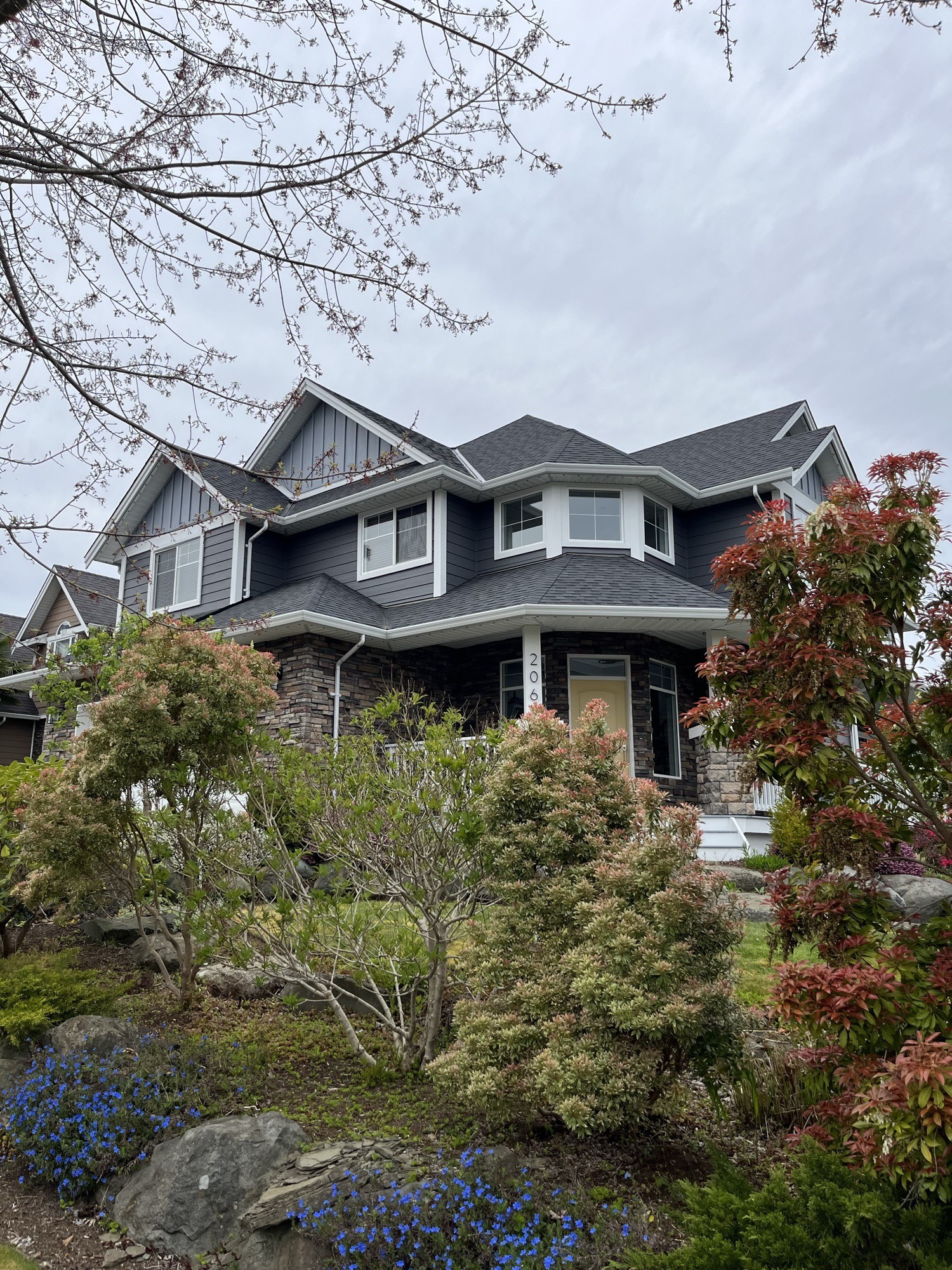 Exterior painting — two-storey home framed by mature trees and spring landscaping, Comox Valley
