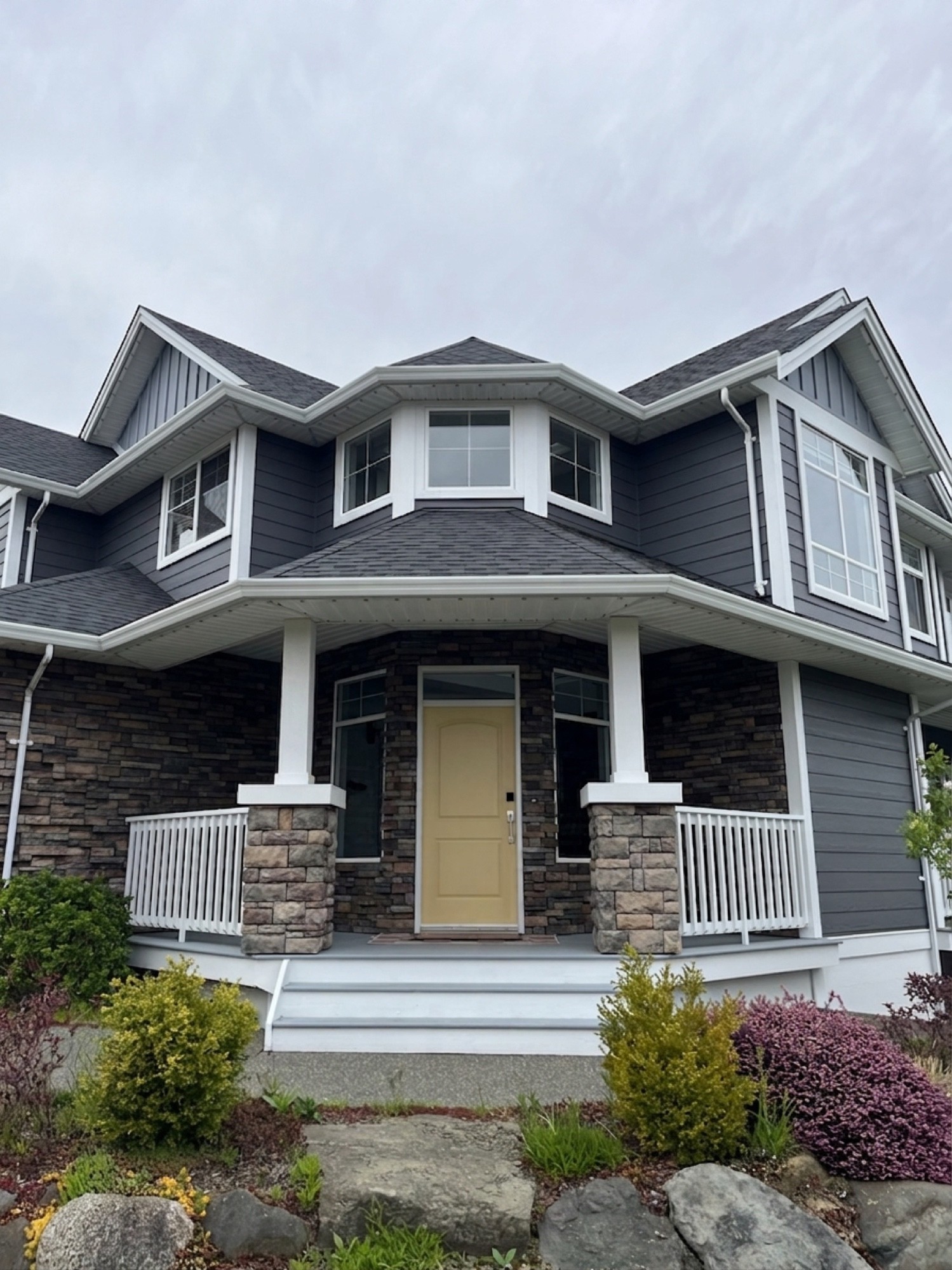 Exterior painting — front of two-storey home with yellow door, dark grey board-and-batten siding, white trim and stone columns
